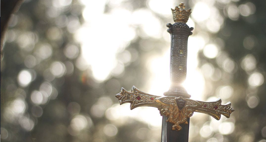 An ornate crowned sword held aloft against sunlit woodland, symbolising empowerment and the will