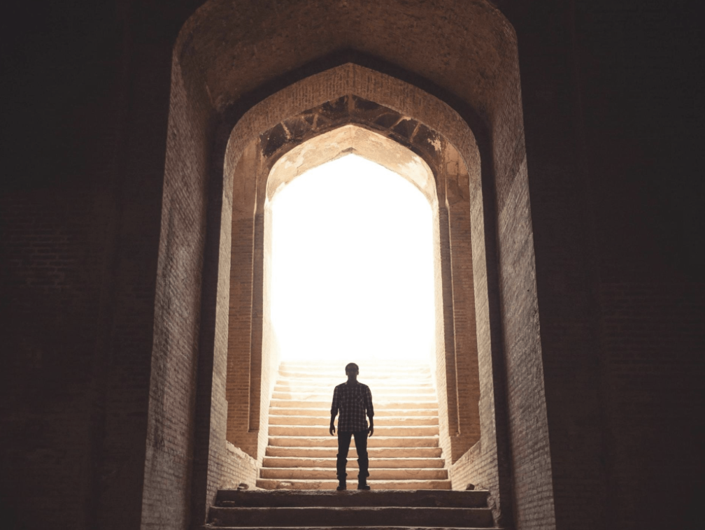 A lone figure standing at the foot of a stone staircase, framed by an ancient archway with bright light beyond