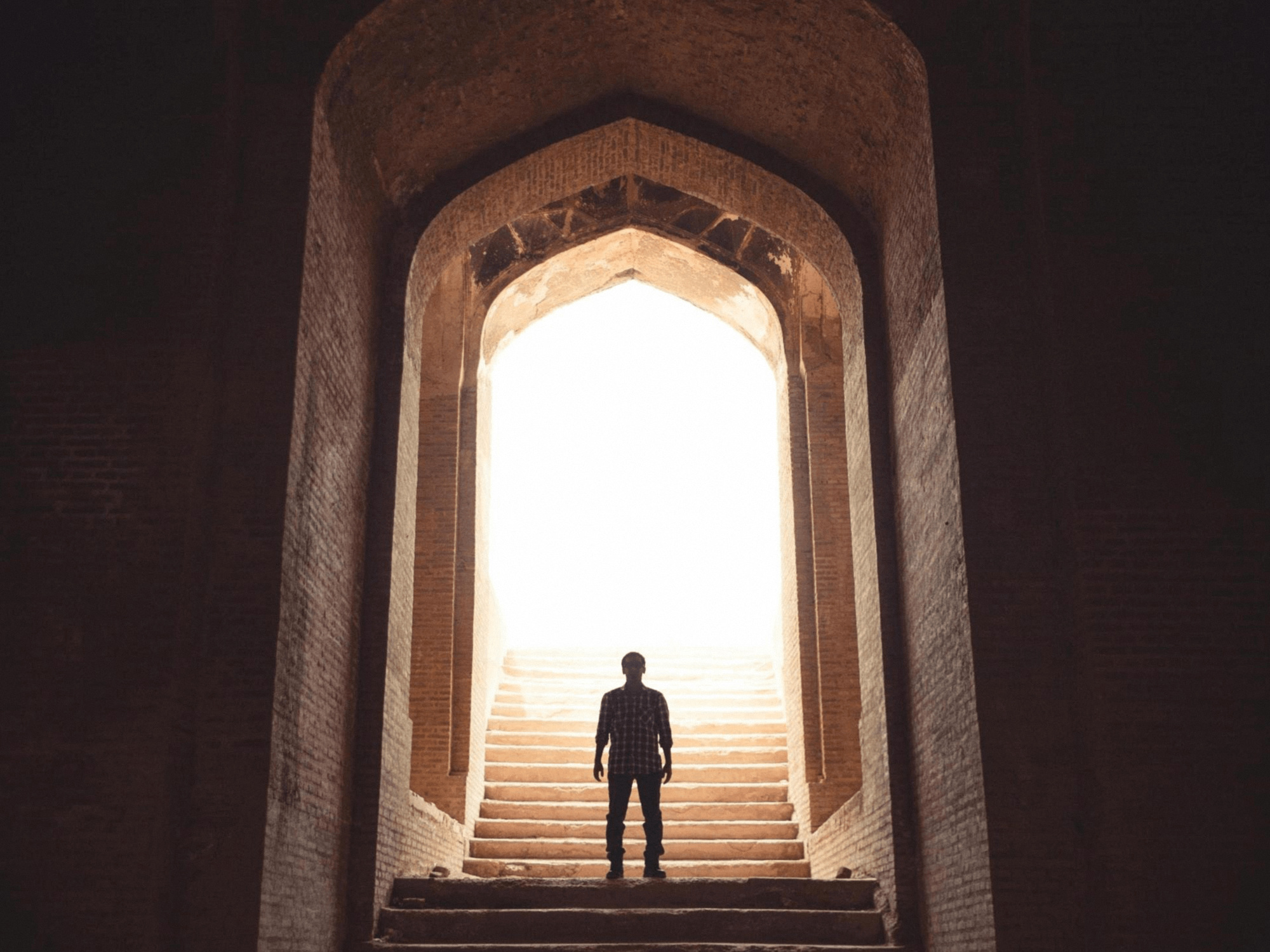 A lone figure standing on ancient stone steps beneath a tall arch, facing a doorway of bright light