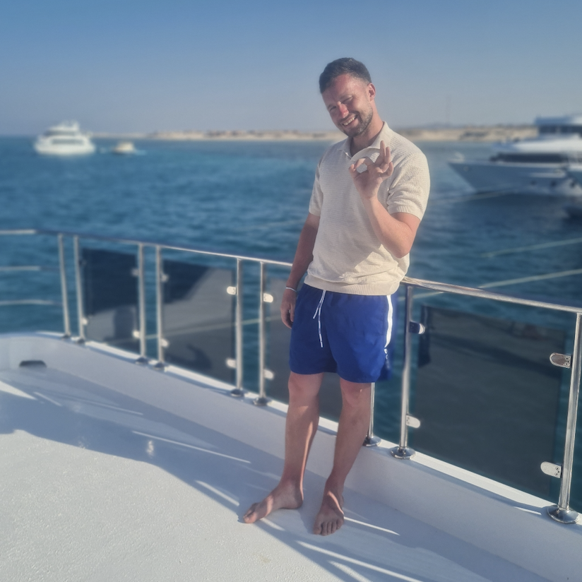 A man standing barefoot on the white deck of a boat, sea and moored yachts behind him